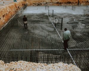 Two workers inspecting a rebar grid foundation at a construction site.